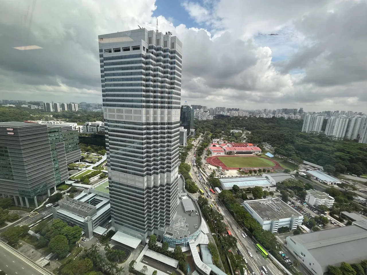 Aerial view of Mapletree Business City with nearby sports field, roads, and greenery under cloudy sky.